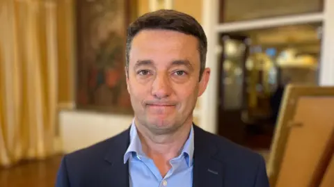 Boley Smillie, a man with black hair looking at the camera. He's wearing a navy suit jacket and a blue button up shirt.