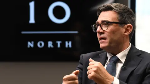 PA Media Andy Burnham is pictured in a suit and glasses, gesturing with both hands while seated. A large screen in the background displays the words “10 NORTH” in white text on a black background.