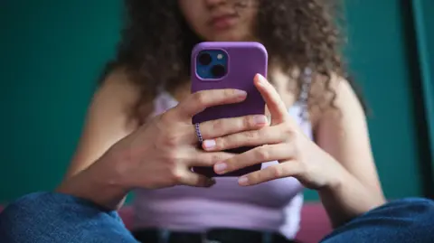 Getty Images A young woman, whose face cannot be seen, sits cross-legged while holding and using her smartphone. She is wearing blue jeans and a lilac vest top.