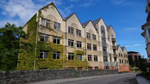BBC Frossard House - The administrative centre for the States of Guernsey. Often nicknamed Custard Castle. On the left side it is covered in green ivy. It is a yellow building with triangular points in five sections.