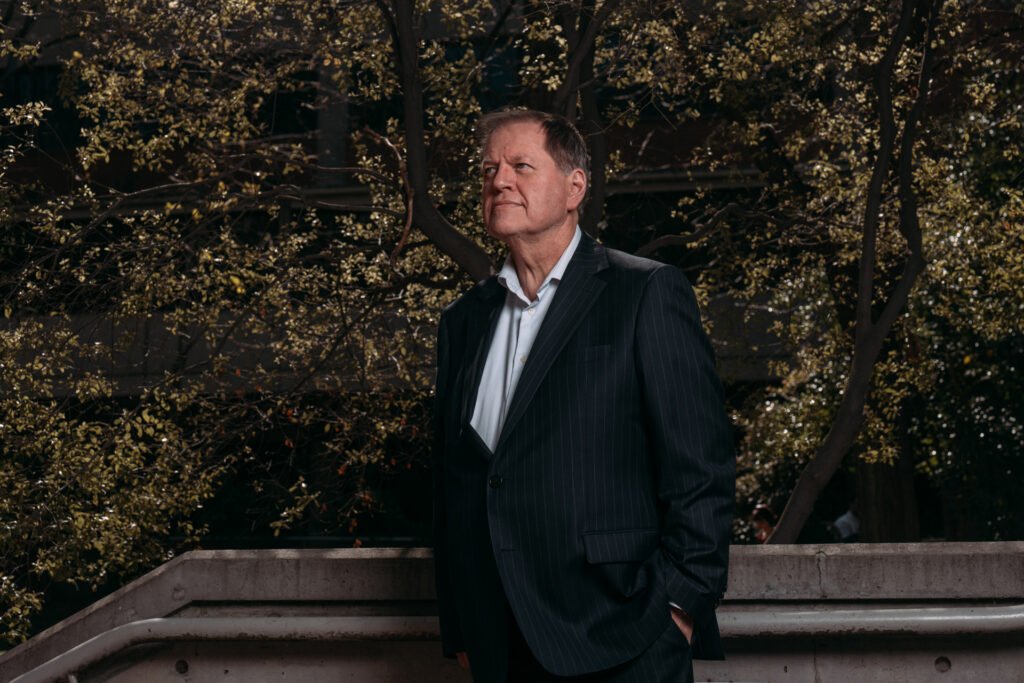 A man in a light shirt and dark blue suit jacket outside in front of a tree.