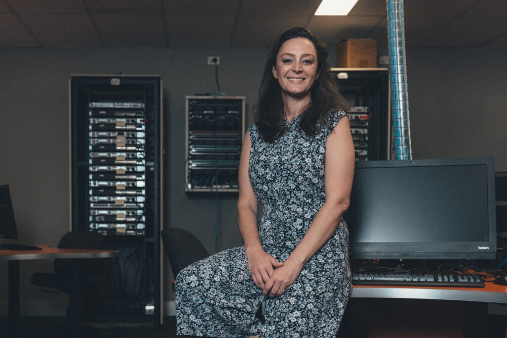 A woman with long brown hair wearing a blue dress stands in a datacenter classroom.