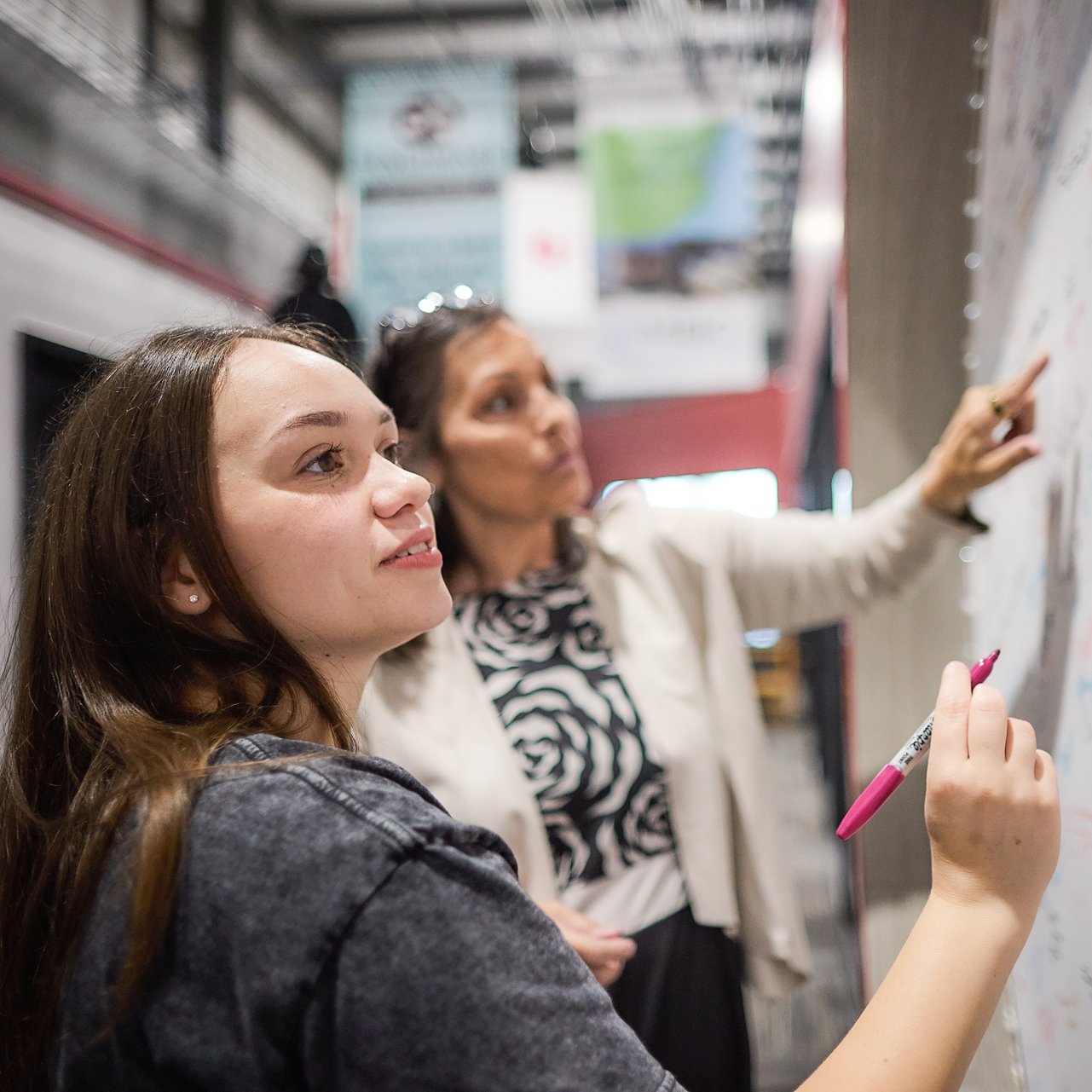 Two dark-haired women, one in a dark shirt and the other in a cream sweater, examine a white board at a teaching lab.