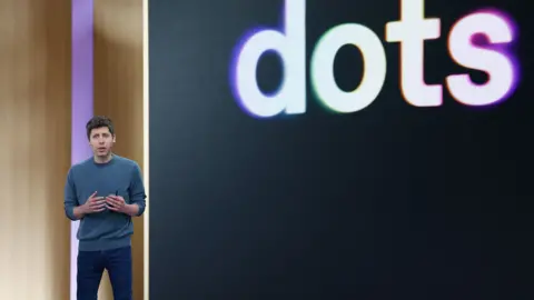 Reuters OpenAI CEO Sam Altman standing on stage at his company's annual developer conference in front of a screen with "dots" displayed behind it.