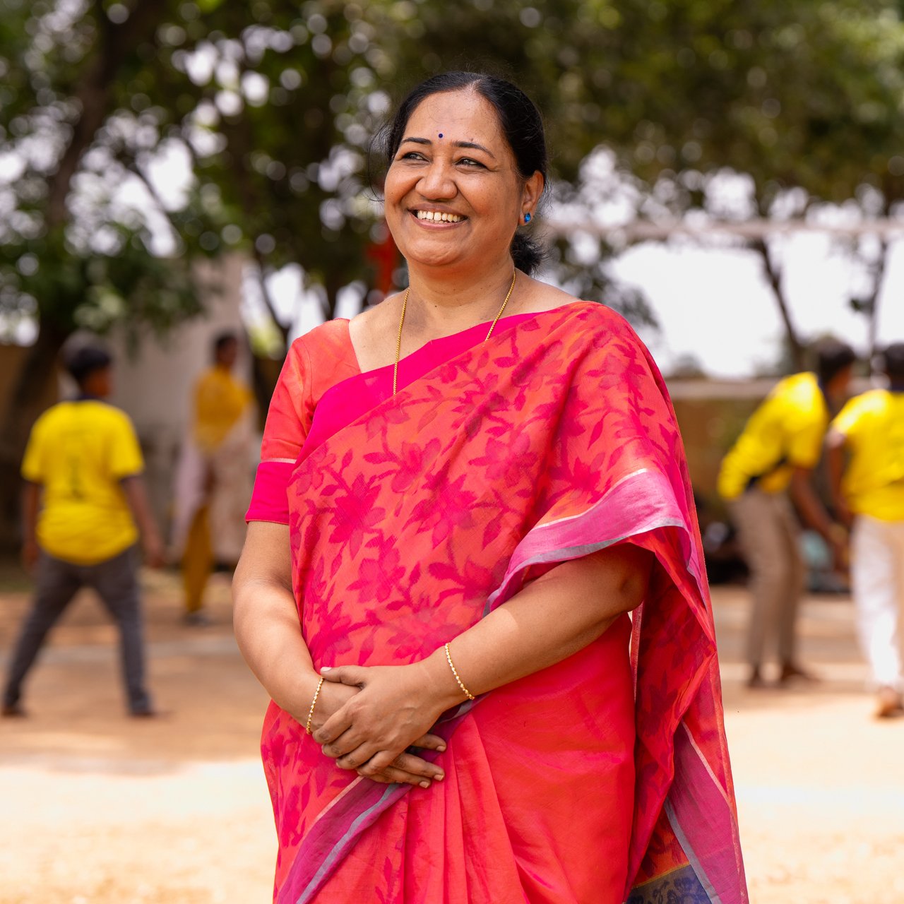 A woman with dark hair wearing a pink sari stands with hands clasped in front of her on a playground.
