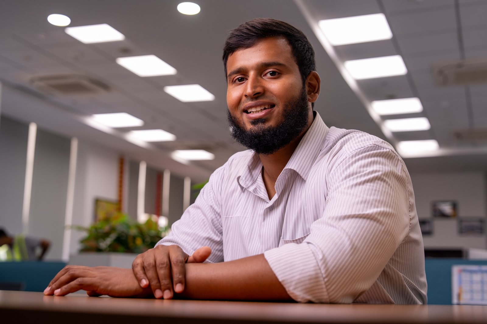 A young man with a beard wearing a white shirt sits at an office desk smiling.