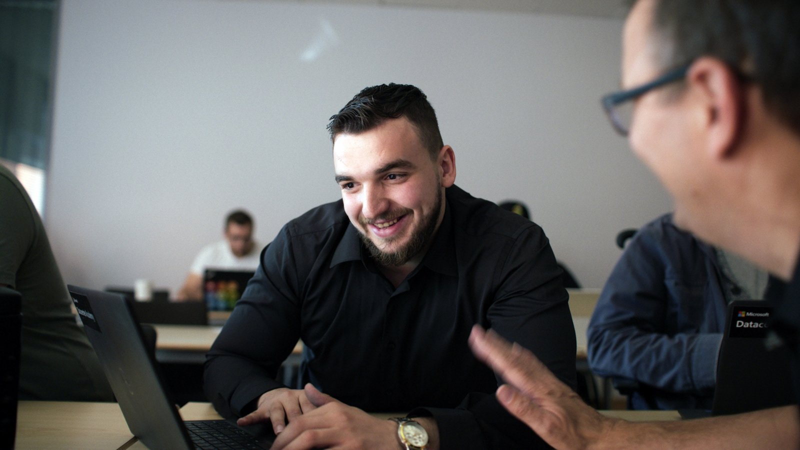 A young man with short dark hair and beard sits at a table talking to another man.