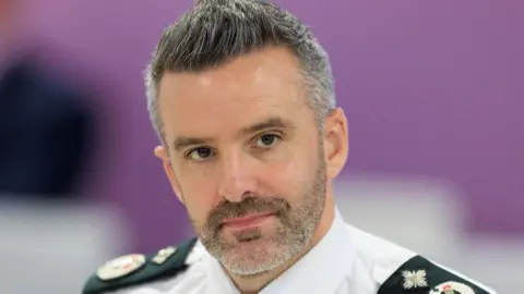 PA Media A man with grey and black hair and beard wearing a white police shirt with black lapels with symbols on them.
