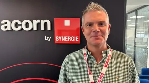 A man with short grey hair smiling at the camera. He has a white and green crosshatched shirt. He has a white and red lanyard around his neck. Next to him is a sign that says Acorn by Synergie.