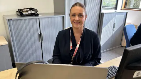 A woman sat behind a desk in an office. She smiles at the camera. She has a dark blue top on and a red and white lanyard.
