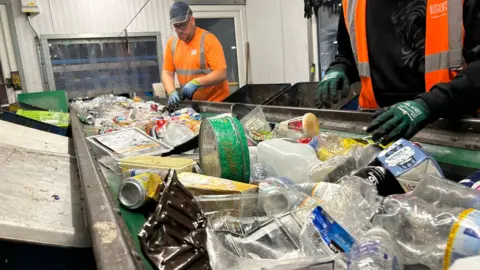 Workers at the Vale of Glamorgan Council's recycling facility in Barry. There is plastic waste on a conveyor belt.